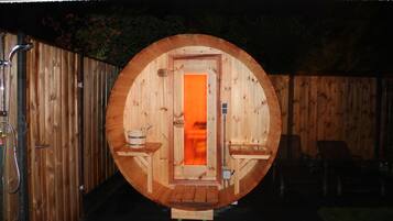 Een houten sauna met een ronde deur, een bank en een kleine tafel met een emmer.