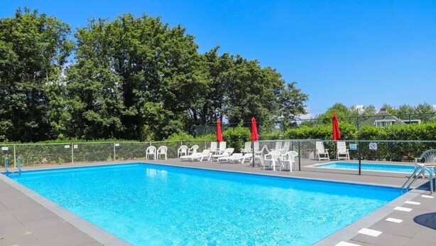 A rectangular swimming pool with clear blue water, surrounded by a fence. There are white lounge chairs and red umbrellas along the poolside.