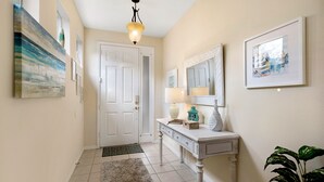 Um corredor com uma porta branca, um console com uma luminária, um quadro emoldurado e uma planta.