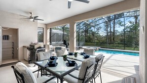 Uma área de refeições ao ar livre espaçosa, com mesa e cadeiras de vidro, ventilador de teto e vista para uma piscina.