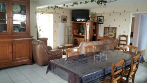 A dining area with a table covered by a protective sheet, wooden chairs, a sofa, and a television on the wall. - 7 km from DERVAL, stone house 4/5 adults + 1 baby. INTERNET BOX. (Conquereuil)