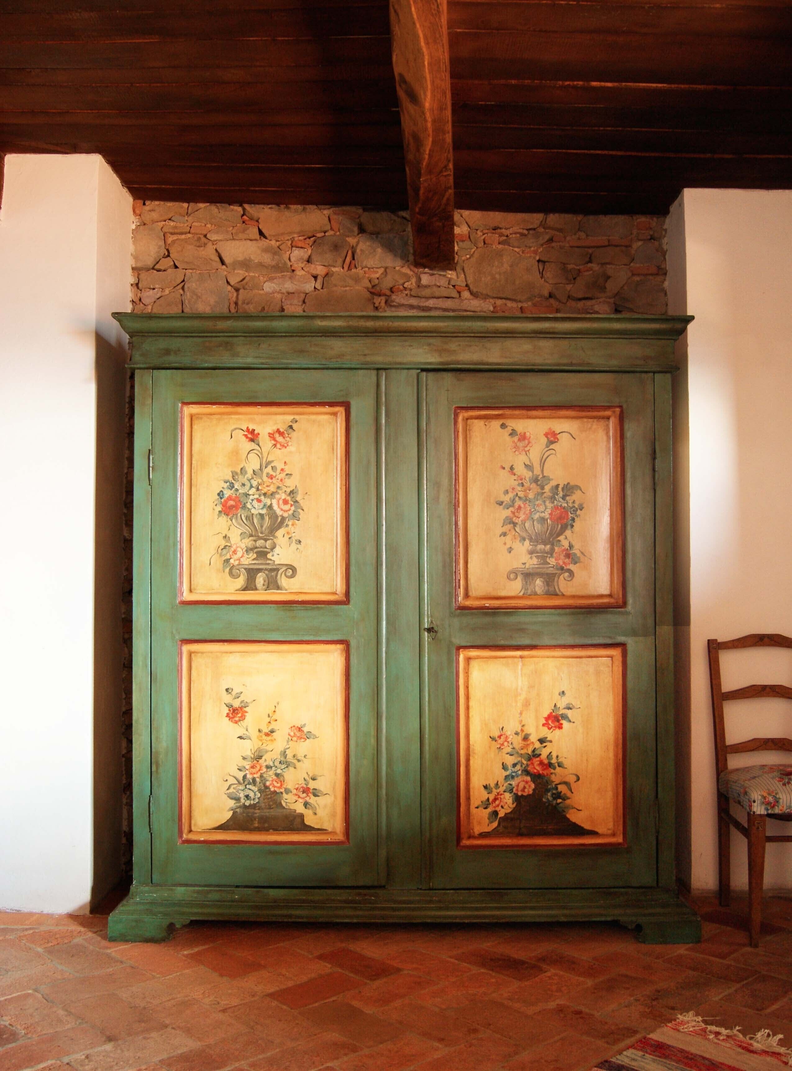 Un armario verde con diseños florales en las puertas, una pared de piedra arriba y una silla de madera a la derecha.