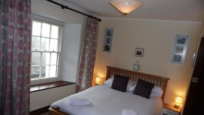 A neatly made bed with white linens and two pillows, a bedside table with a lamp, a window with curtains, and a wall with framed pictures.