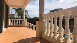 Un balcón techado con balaustradas blancas, piso de baldosas y vista a palmeras y un edificio al fondo.