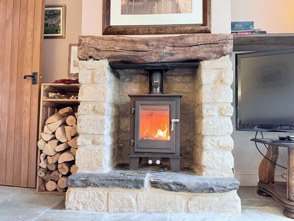 A stone fireplace with a wood-burning stove, a stack of firewood to the left, and a television on a wooden stand to the right. - 4 Star Cotswold Stone Cottage (Winchcombe)