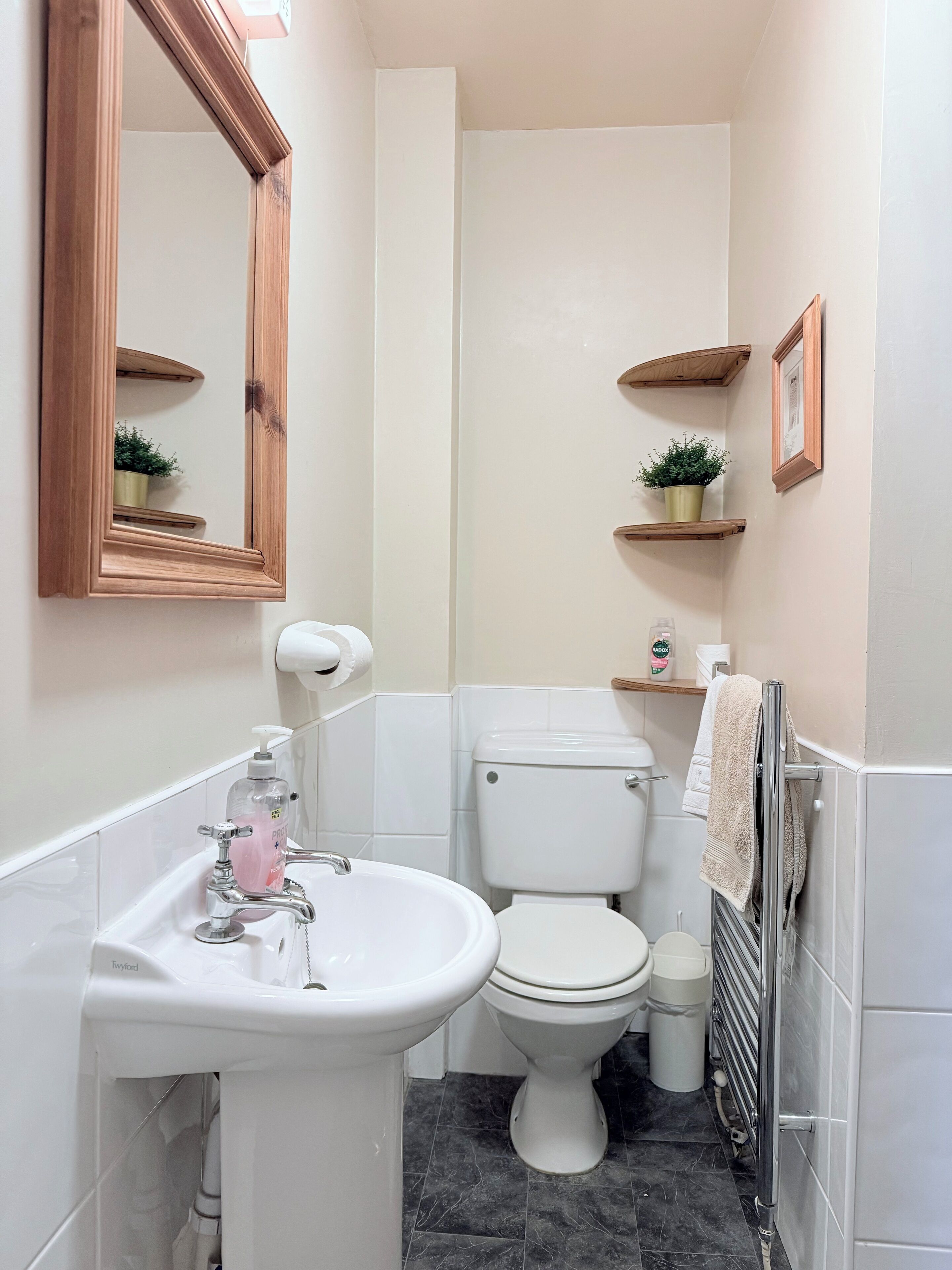 Ein Badezimmer mit einem weißen Waschbecken, einer Toilette und einem Spiegel. Holzregale mit Pflanzen und einem Handtuchhalter sind ebenfalls vorhanden.