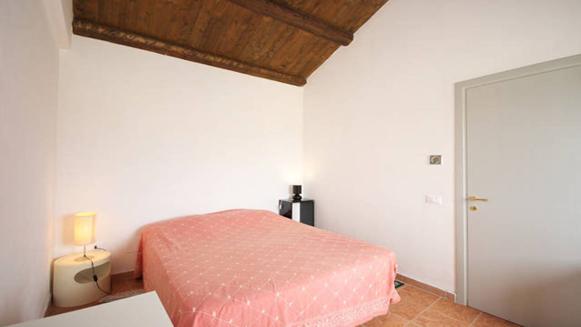A bedroom with a bed, a nightstand, a lamp, a door, and a ceiling with exposed beams.