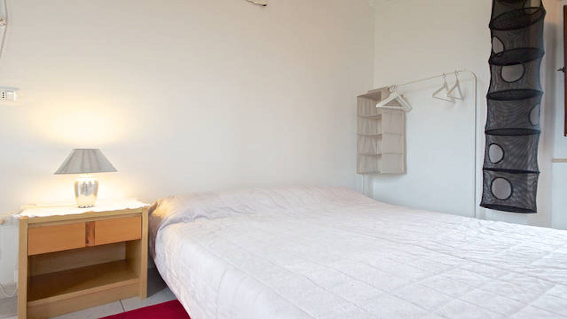 Ein Schlafzimmer mit Bett, Nachttisch mit Lampe und Kleiderschrank mit Kleidung.