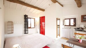 Ein Schlafzimmer mit einem Bett, einer hölzernen Bank, einer roten Tür und einer Küchenzeile mit Spüle und Schränken.