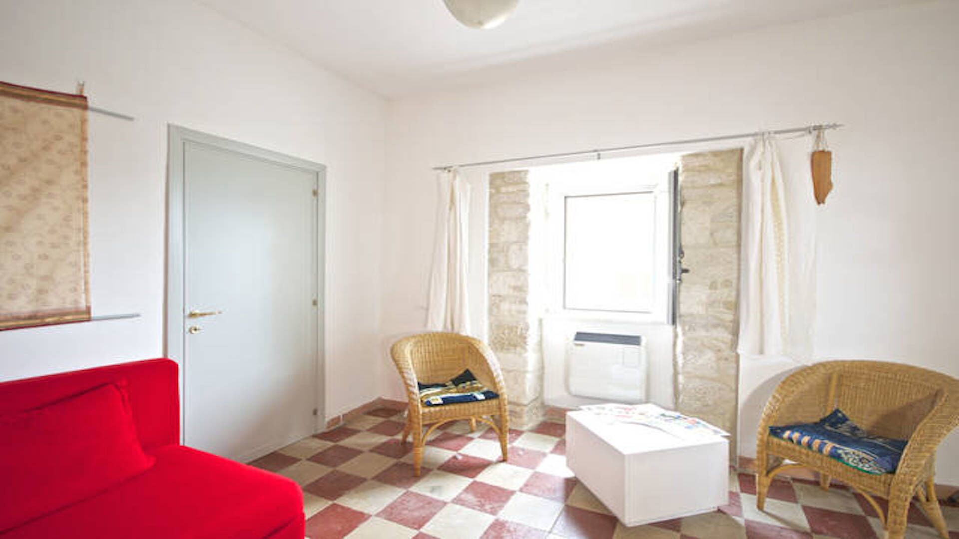 Een kamer met een rode bank, twee rieten stoelen, een witte tafel en een zwart-wit betegelde vloer.