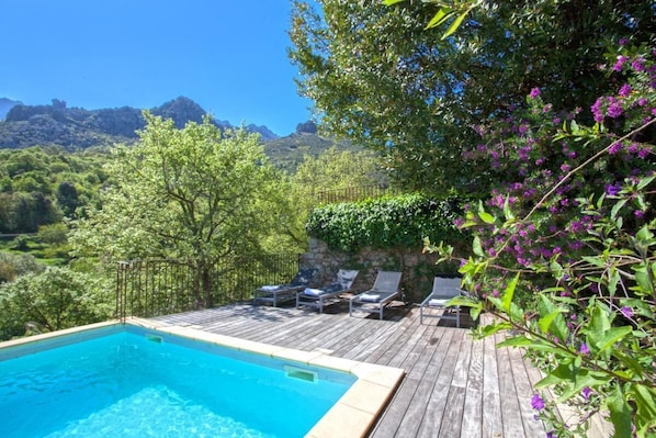 Une piscine dotée d’une terrasse en bois et de chaises longues, entourée de verdure et avec des montagnes en toile de fond.