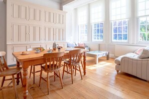 A spacious room with a wooden dining table, chairs, and a sofa. There are large windows providing natural light and a view of the outdoors.