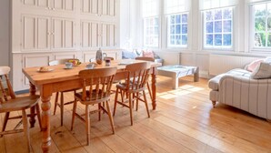 Een ruime kamer met een houten eettafel, stoelen en een bank. Grote ramen zorgen voor natuurlijk licht en uitzicht op het groen.