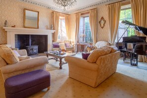 A cozy living room with a fireplace, comfortable seating, a piano, and large windows with curtains.