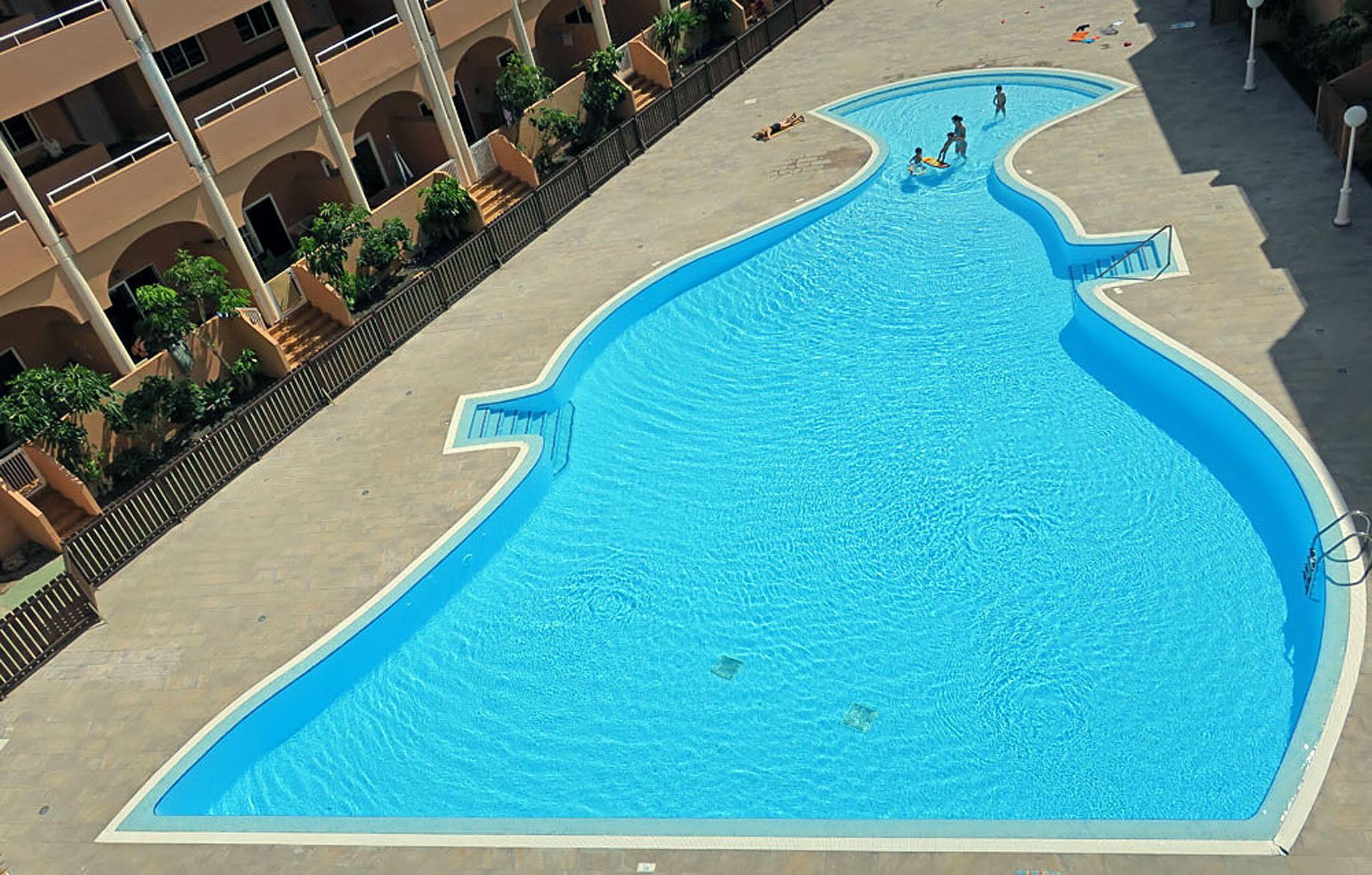 Una piscina grande y de forma peculiar con superficie de agua azul cristalina, rodeada por una cerca baja y un edificio con varios balcones.