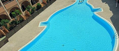 Una piscina grande y de forma peculiar con superficie de agua azul cristalina, rodeada por una cerca baja y un edificio con varios balcones.