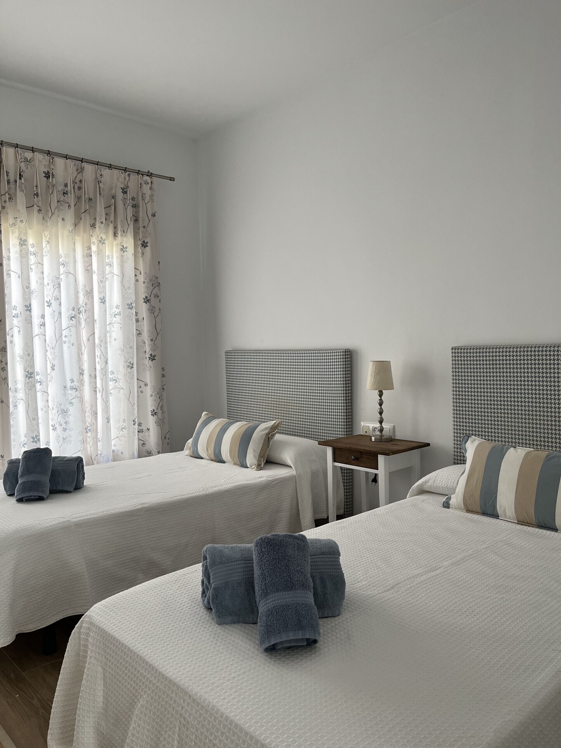 A hotel room with two beds, a bedside table with a lamp, and a window with floral curtains.