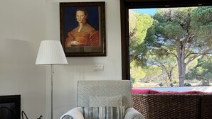 Un salón con un sillón estampado, un cuadro en la pared, una lámpara y vistas a árboles a través de un ventanal grande.
