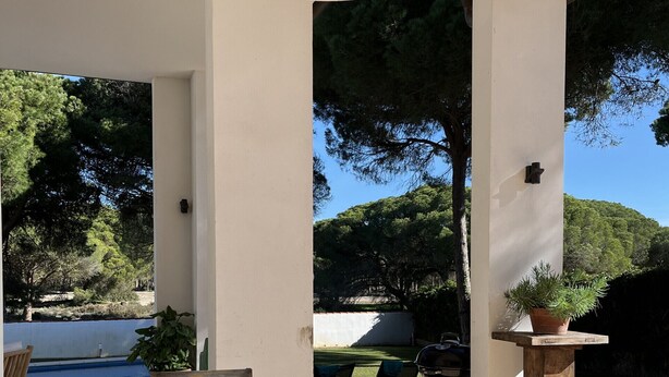 Un área de comedor al aire libre con una mesa de madera dispuesta para seis personas, rodeada de sillas de mimbre, un techo blanco y vistas a una piscina y árboles.