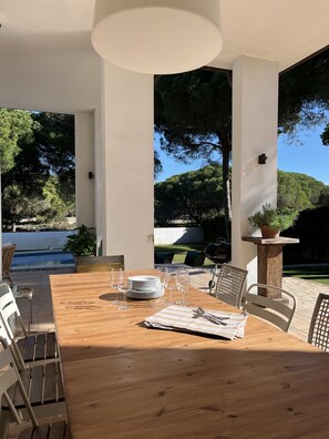 An outdoor dining area with a wooden table set for six, surrounded by wicker chairs, a white ceiling, and a view of a swimming pool and trees.
