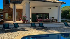 Una casa moderna con fachada de piedra, un porche cubierto con sillones de descanso y una piscina.