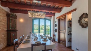 A dining room with a long table set for a meal, a chandelier, and a view of the countryside through large windows. - Casale Vigneto - 3 bedrooms, pool, AC, wifi, pets, Marche (Mogliano)