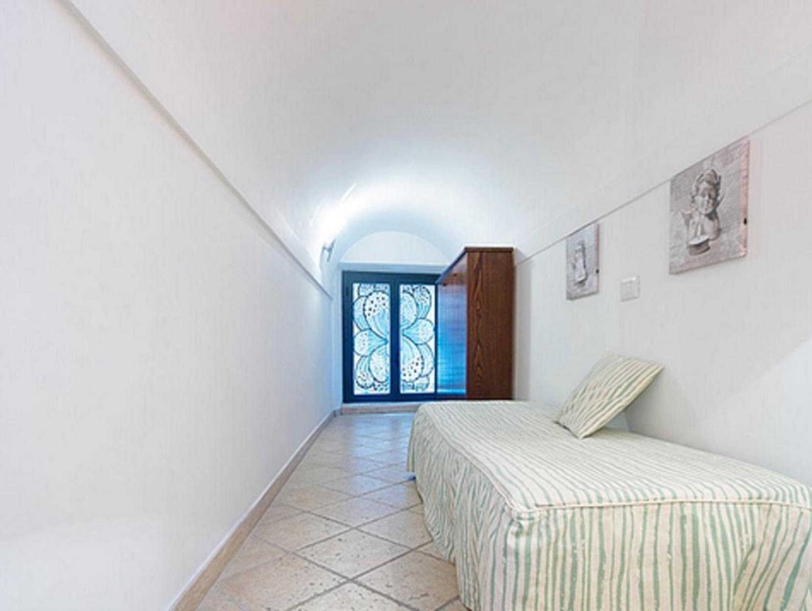 Un pasillo estrecho con una cama a la derecha, un armario de madera y una puerta con un panel de vidrio azul.