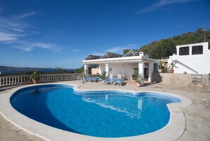 A swimming pool with a curved edge, surrounded by lounge chairs and potted plants, with a view of the sea and a white building in the background.