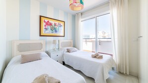Ein Hotelzimmer mit zwei Betten, einer farbenfrohen Hängelampe, einem gerahmten Blumenbild und einem Balkon mit Vorhängen.