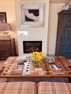 A living room with a fireplace, a table with magazines, a vase of flowers, and a framed picture on the wall.
