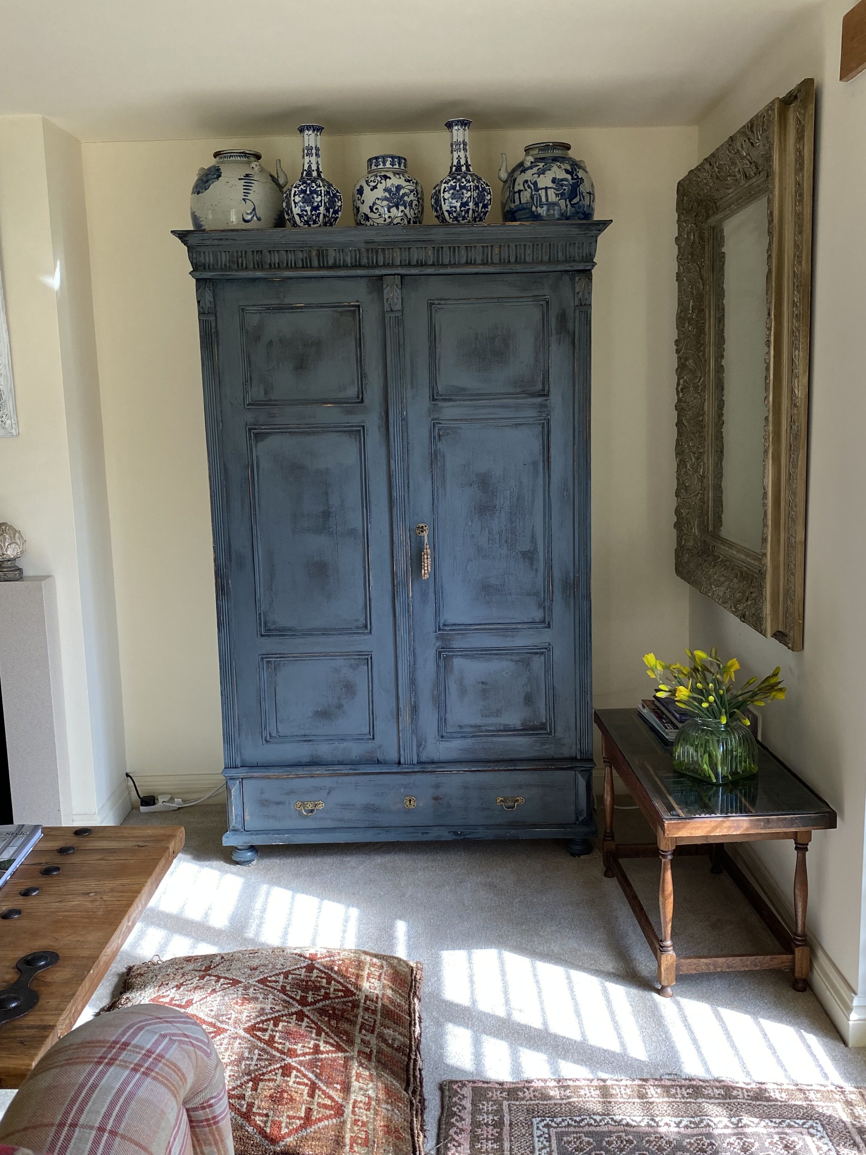 A blue armoire with decorative vases on top, a mirror, and a small table with a vase of flowers.
