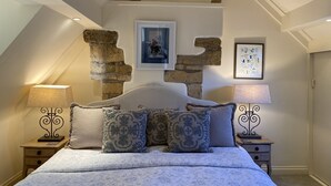 Un dormitorio con pared de piedra, una cama con ropa de cama estampada, dos mesitas de noche con lámparas y un cuadro enmarcado en la pared.