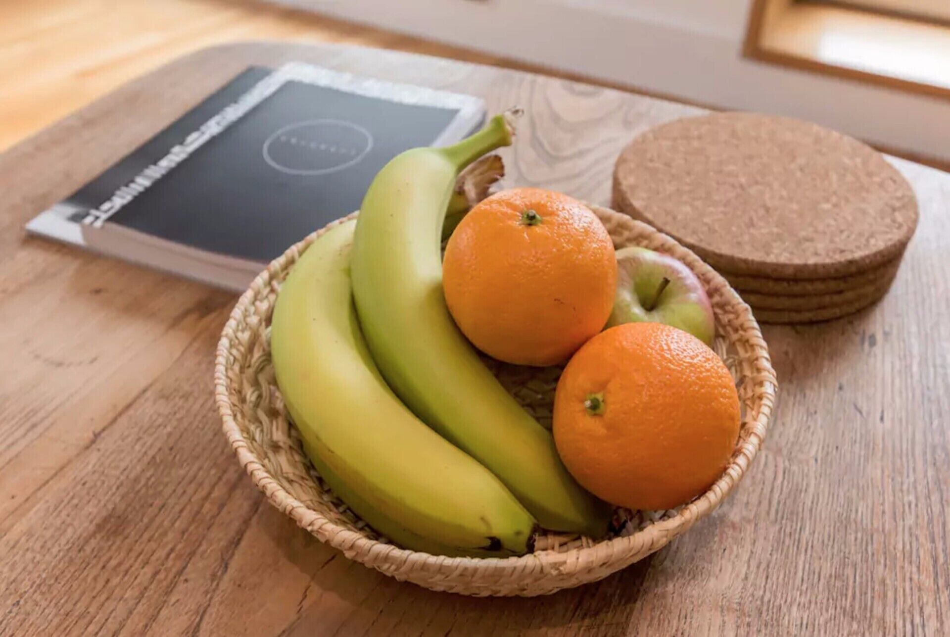 A wicker basket containing bananas and oranges on a wooden table, with a book and coasters in the background.