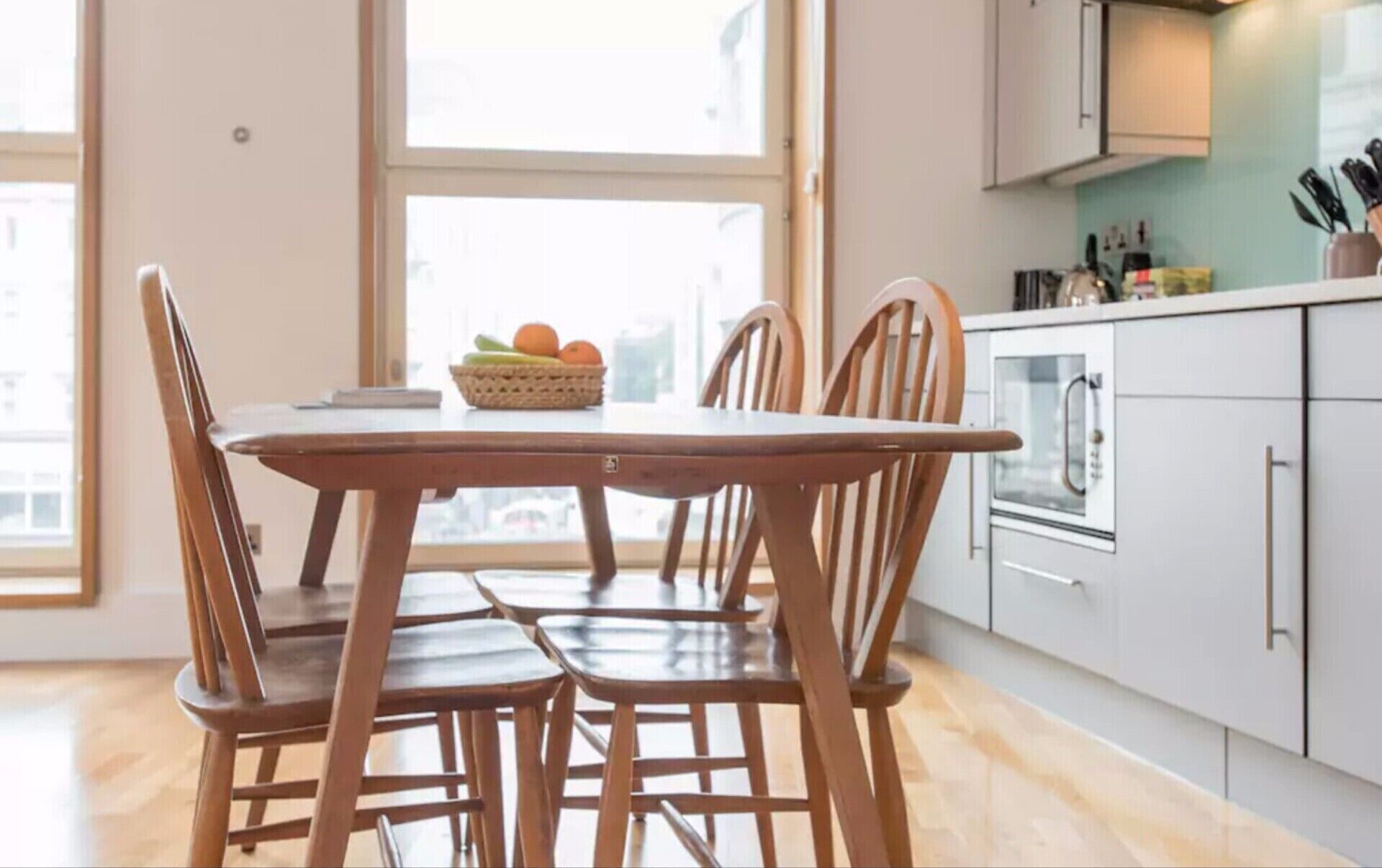 Ein Essbereich mit einem Holztisch und Stühlen, einer Obstkorb und Blick auf eine Küche mit Schränken und einem Backofen.