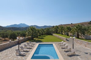 Une piscine rectangulaire bordée de chaises longues et d’un mur en pierre, entourée de palmiers et offrant une vue sur les montagnes.
