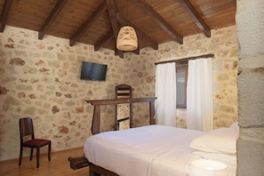 Une chambre à coucher avec un plafond en bois, des murs en pierre, un lit, une chaise, une table, un téléviseur et une fenêtre avec des rideaux.