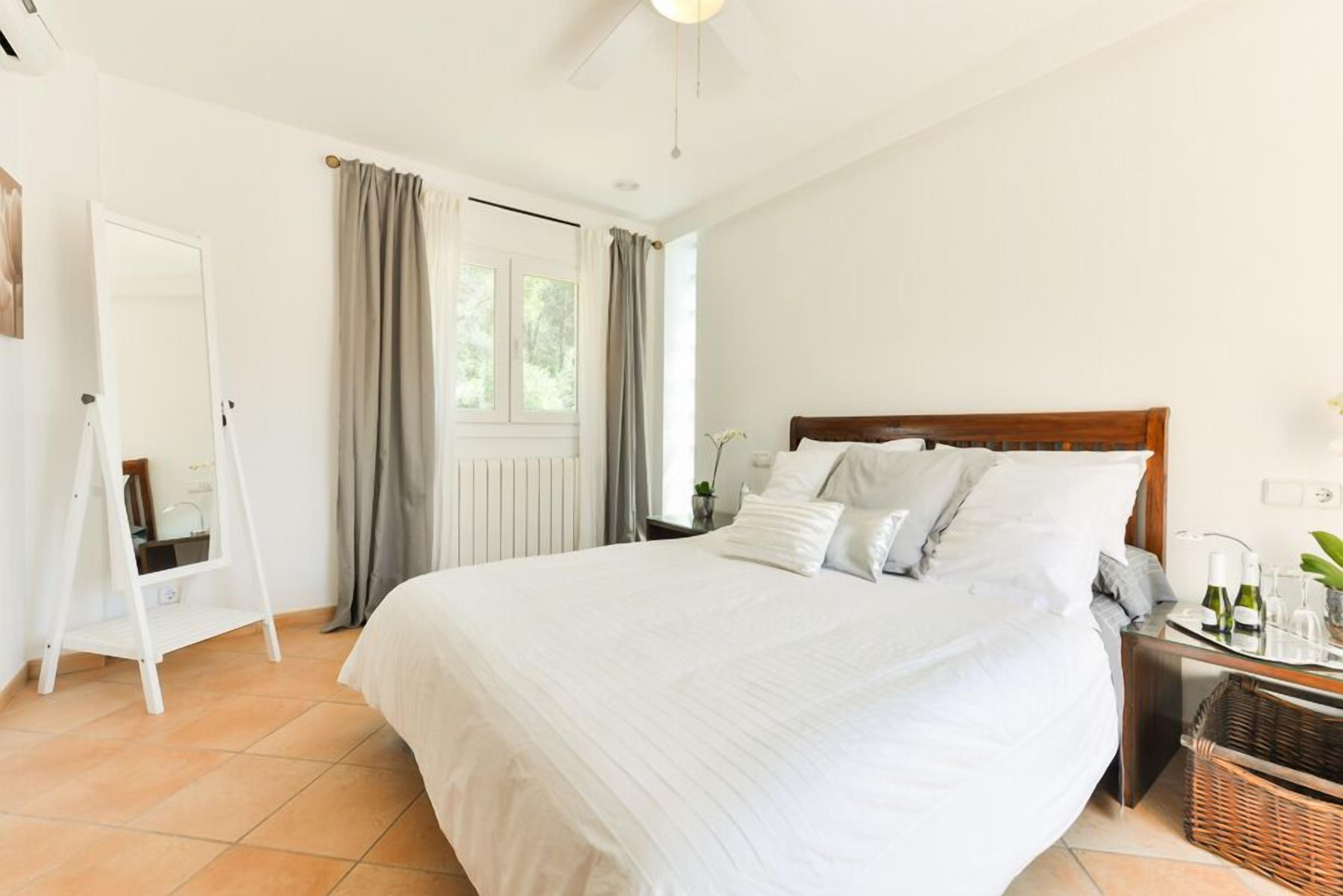 Ein Schlafzimmer mit einem großen Bett, einem Nachttisch, einem Spiegel, einem Fenster mit Vorhängen und einem Deckenventilator.