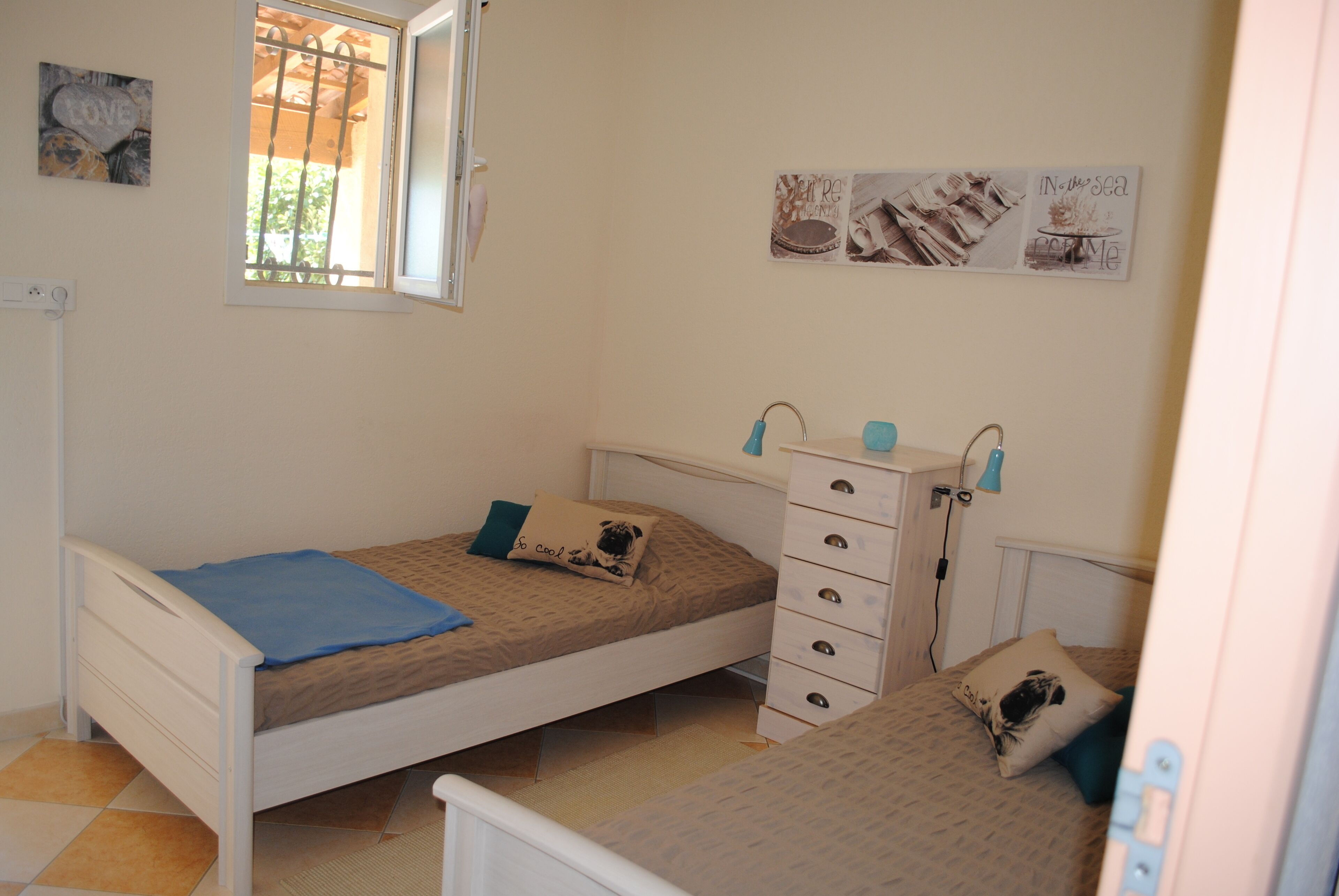 Une chambre avec deux lits simples, une commode, une fenêtre et des éléments décoratifs sur les murs.