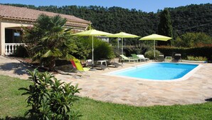 A swimming pool with stone tiles, surrounded by lounge chairs and umbrellas, with a house and forested hill in the background.