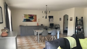 A living room with a grey sofa, a dining table with chairs, a kitchen area with grey cabinets, and a chandelier.