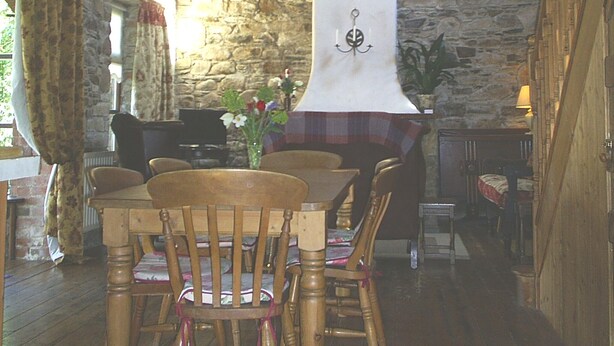 A rustic dining area with wooden chairs and a table, stone walls, a hanging chandelier, and a clock on the wall.