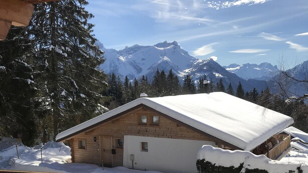 Uma cabana de madeira com telhado coberto de neve, rodeada por neve e árvores, com uma cordilheira ao fundo.