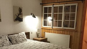 Quarto com cabeceira de madeira, cama com roupa de cama floral, mesa de cabeceira com abajur, decoração de cabeça de veado fixada na parede e janela com vista para o exterior.
