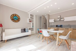 A modern living room with a white sofa, a dining table with white chairs, a kitchen area with wooden cabinets, and a wall clock. - Casa La Rosa dei Venti (Macari)