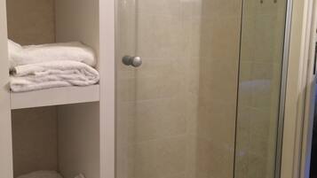 Ducha envuelta en vidrio con pared de azulejos blancos y repisa con toallas dobladas.