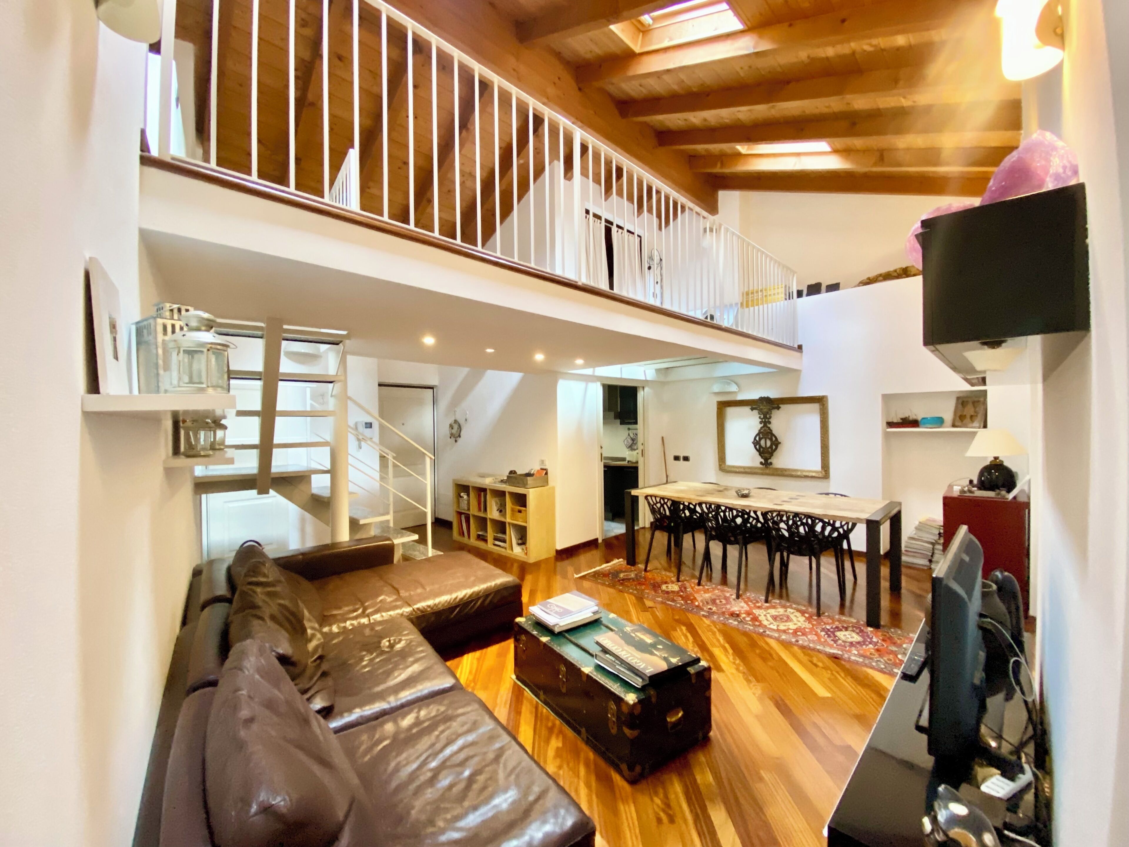 A spacious living area with a high ceiling, a staircase leading to an upper level, a dining area with a table and chairs, and a seating area with a sofa and a coffee table.