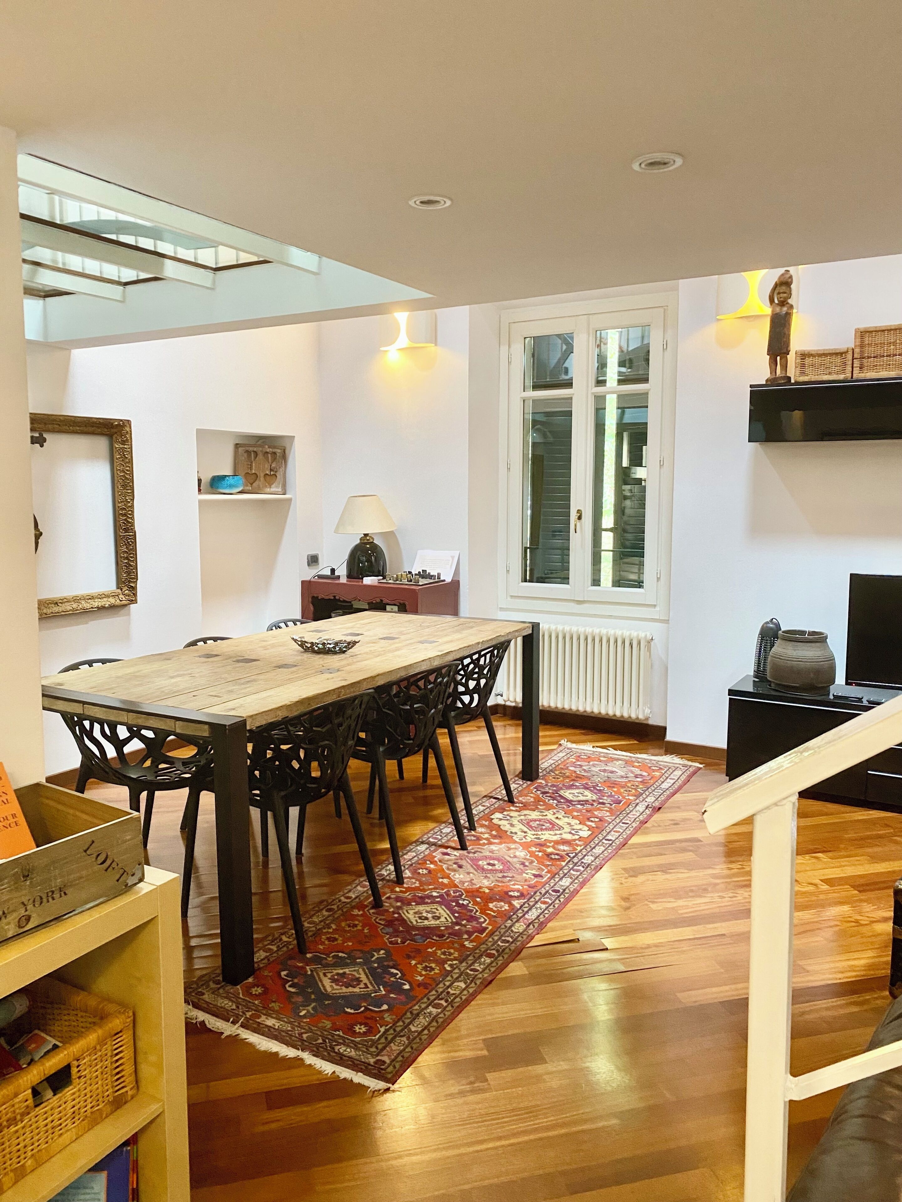 A dining area with a wooden table, black chairs, a patterned rug, and a window with blinds.