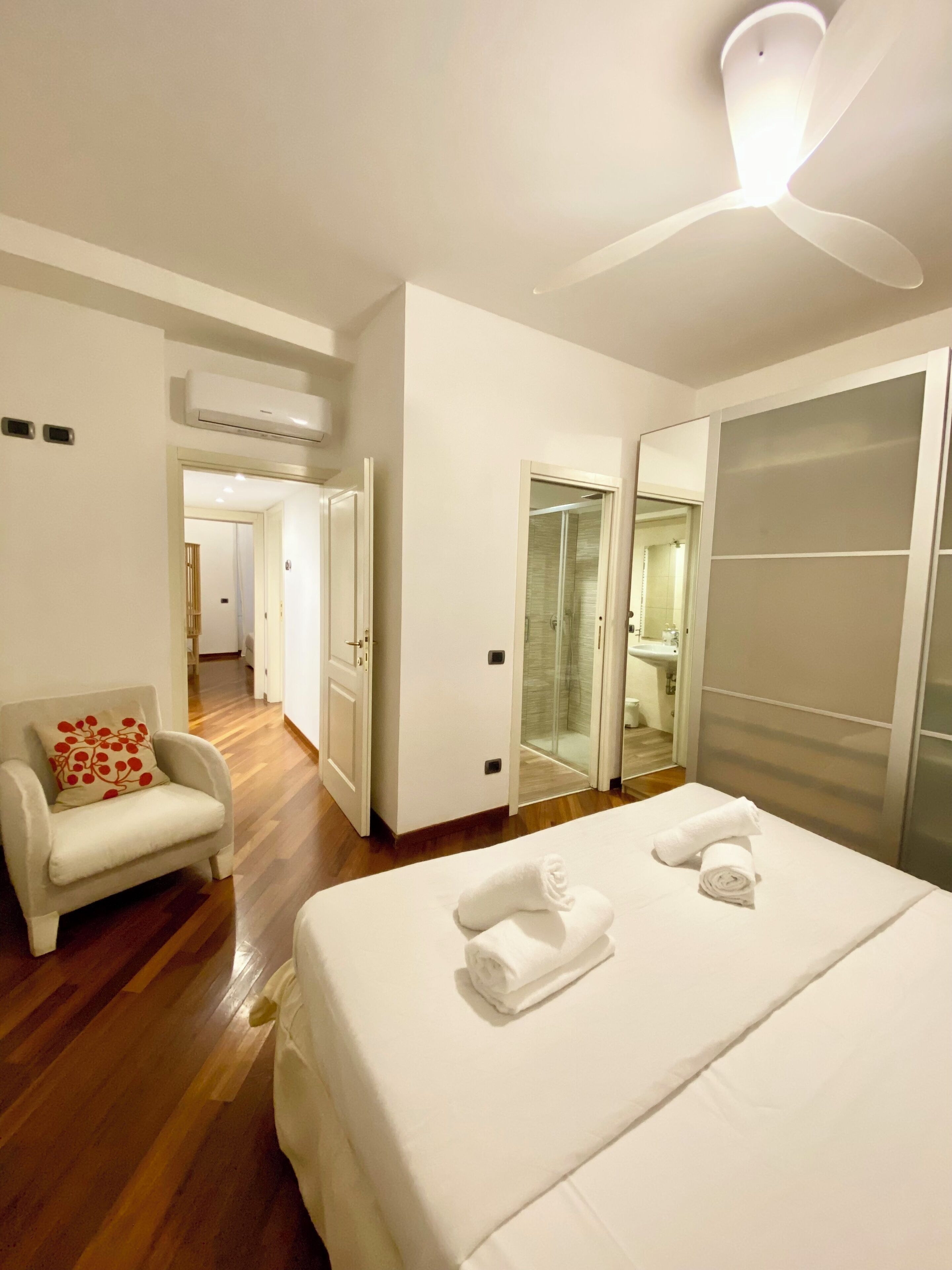 Une chambre d’hôtel avec un lit, une chaise, un ventilateur de plafond et une salle de bain visible à travers une porte ouverte.