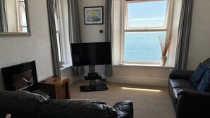 Ein Wohnzimmer mit großem Flachbildfernseher, einem Holztisch, einem schwarzen Ledersofa und Meerblick durch ein großes Fenster.