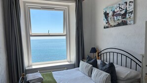 Ein Schlafzimmer mit großem Fenster, einem Bett mit weißen Leinen, einer grünen Tagesdecke und einem Gemälde an der Wand.